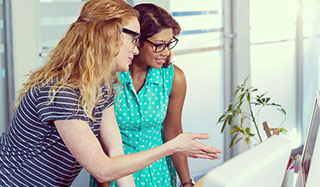 Two business woman standing talking next to computer screen